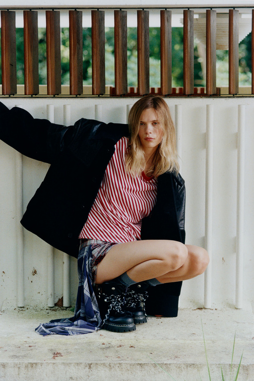 A woman with blonde hair wearing a black jacket, striped shirt, and black boots crouches in front of a white wall with a wood railing.