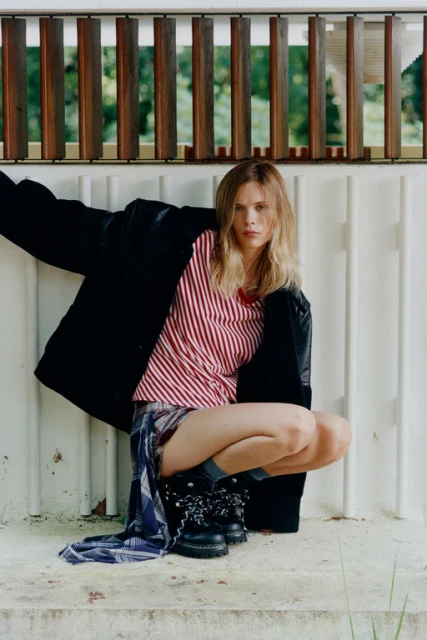 A woman with blonde hair wearing a black jacket, striped shirt, and black boots crouches in front of a white wall with a wood railing.