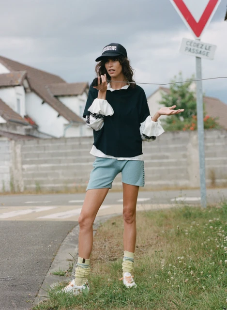A woman in fashionable streetwear is holding a wire near a road sign on an overcast day. She's wearing a cap, black top, and blue shorts.