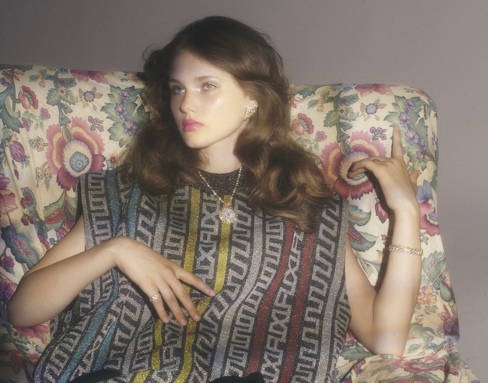 Portrait of a young adult female wearing a patterned top and jewelry, reclining on a floral armchair in a warm-toned indoor setting.