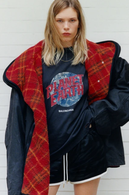 A blonde woman poses in a navy Planet Earth tee, shiny black shorts, and a plaid-lined jacket against a neutral backdrop.