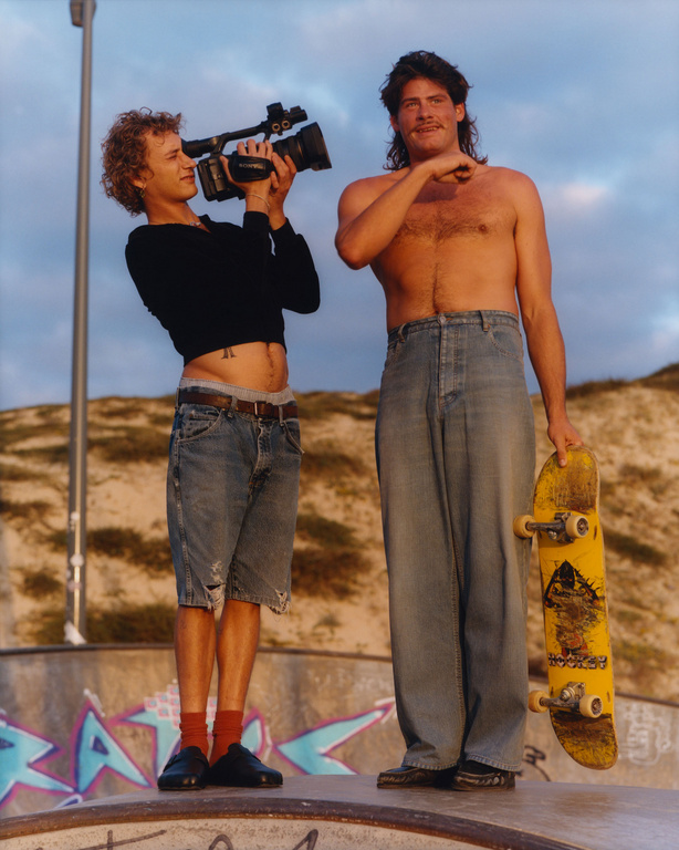 Two men at a skatepark: one films with a vintage camera, the other holds a skateboard, showcasing 90s-inspired fashion and outdoor activity.