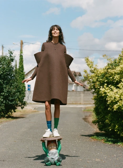 Fashion shot of a woman in a brown dress standing on a garden gnome statue on a street on a sunny day with blue sky and green foliage.