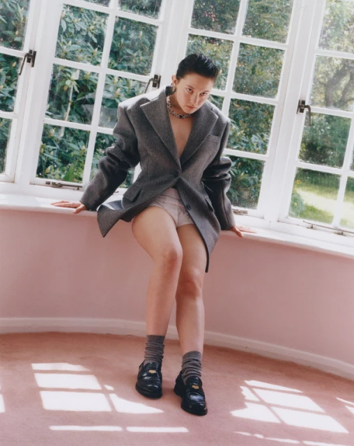 A woman in an oversized grey blazer sits on a windowsill with a garden in the background. She wears socks and loafers and a chunky silver necklace.