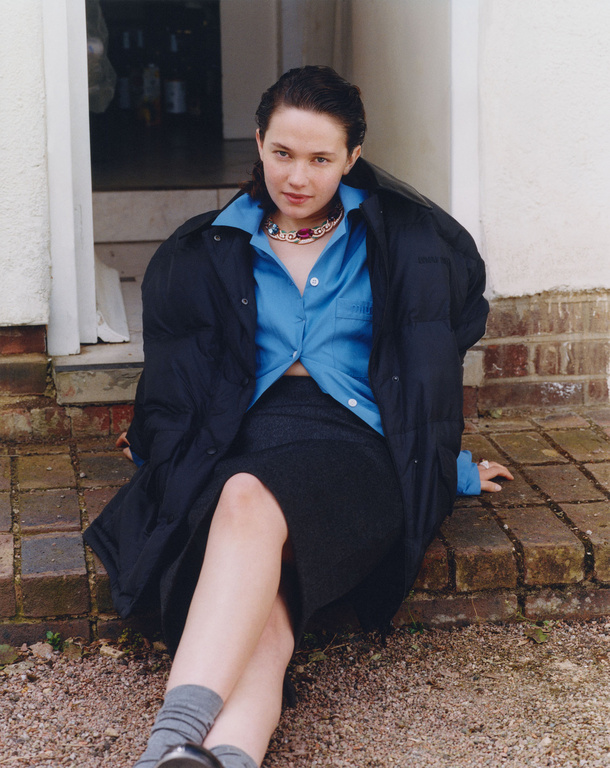 A woman in a blue button-down shirt and dark skirt, wearing a puffer jacket, sits on a brick surface outdoors in a casual pose.