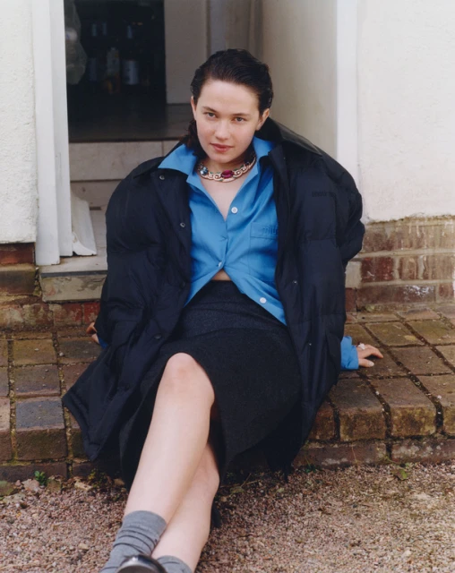 A woman in a blue button-down shirt and dark skirt, wearing a puffer jacket, sits on a brick surface outdoors in a casual pose.