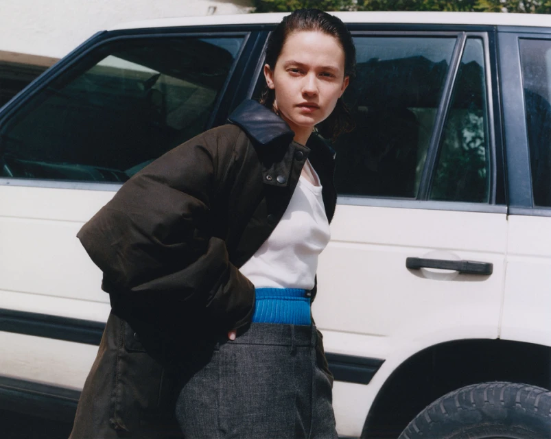 Woman in dark jacket stands near a white SUV outdoors. She wears a white top and grey pants in natural light, showcasing casual fashion.