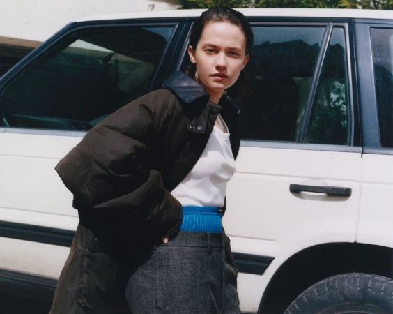 Woman in dark jacket stands near a white SUV outdoors. She wears a white top and grey pants in natural light, showcasing casual fashion.
