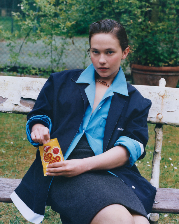 A woman seated outdoors on a bench holding a yellow snack packet, wearing a blue shirt and dark jacket styled by Helga Burrill