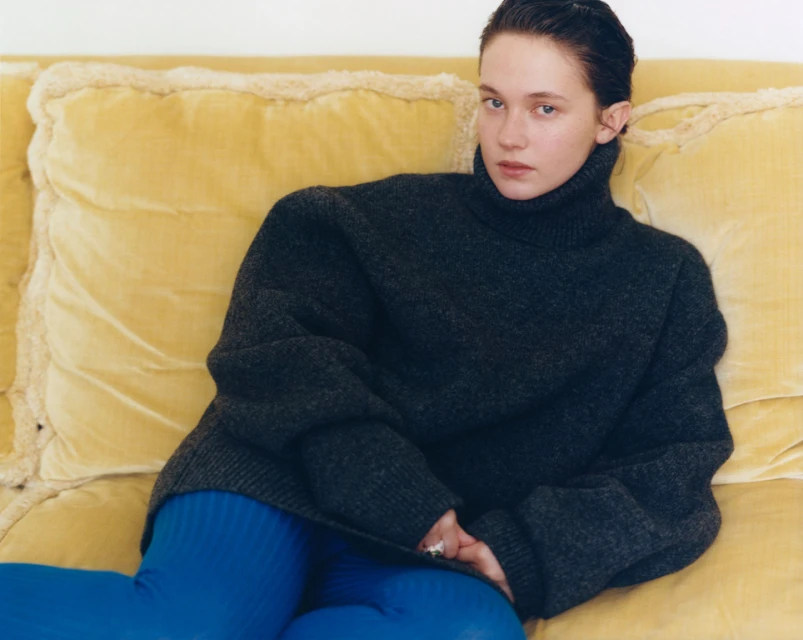 A fair-skinned young woman with dark hair poses on a yellow tufted sofa, wearing a gray turtleneck sweater and blue pants in a studio shot.