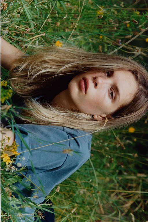 Woman with long blonde hair lies in tall grass and yellow flowers, wearing a blue shirt, photographed outdoors with a shallow depth of field.