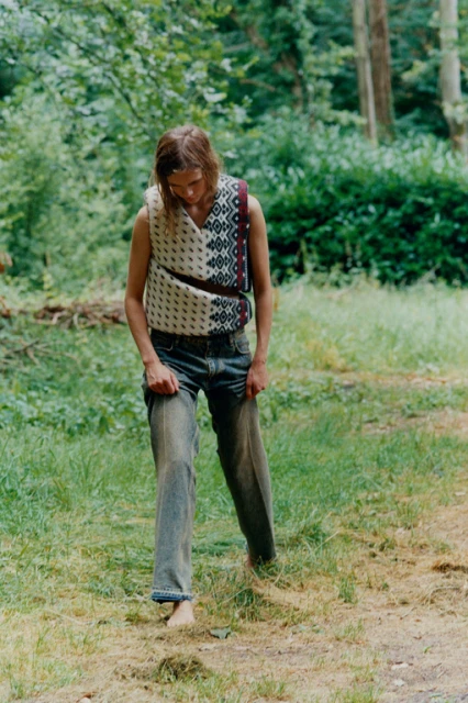 A person stands barefoot in a grassy clearing wearing jeans and a patterned vest, with a forest visible in the blurred background.