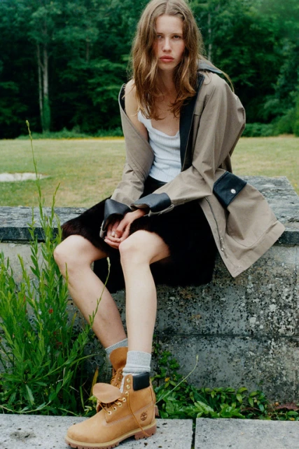 Young woman in tan work boots sits on stone in a park, wearing a white tank top, black skirt, and khaki trench coat.