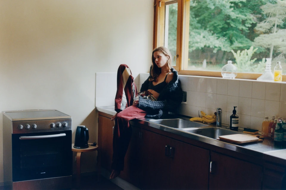 A young woman sits on a kitchen counter next to a window, wearing a black top and cardigan, with cooking appliances in the background.