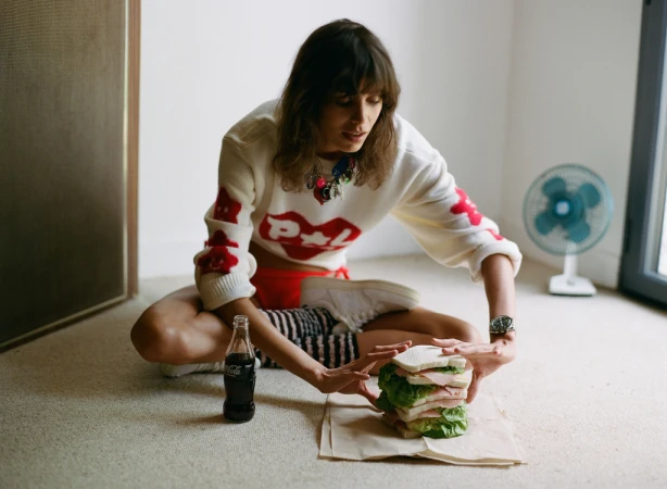 Woman sitting on the floor interacting with a large stacked sandwich and a glass soda bottle, styled by Helga Burrill