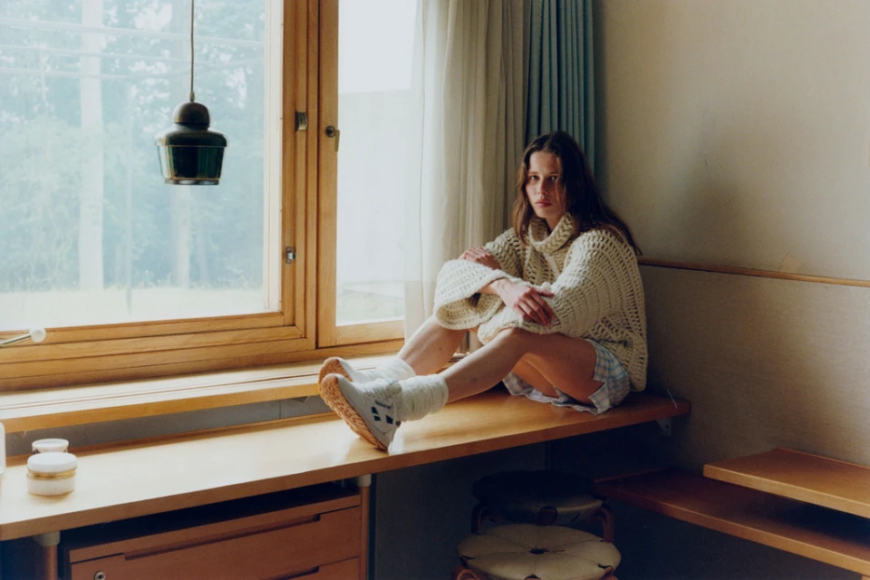 A young woman in a knitted sweater sits on a windowsill with natural light, creating a soft, cozy indoor scene with neutral tones.