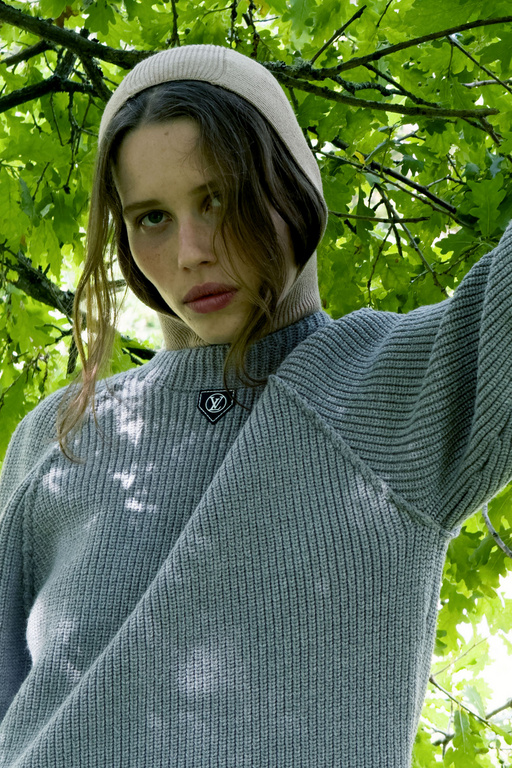 Portrait of a young woman wearing a gray knit sweater and beige beanie against a green leafy background. Fashion editorial style.