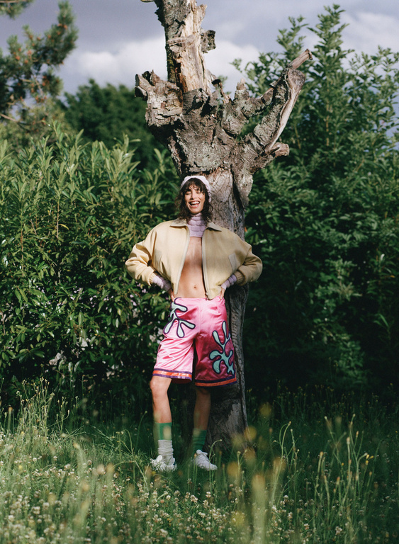 Adult posing in pink patterned shorts and tan jacket next to a tree in a grassy field with green trees and cloudy skies.
