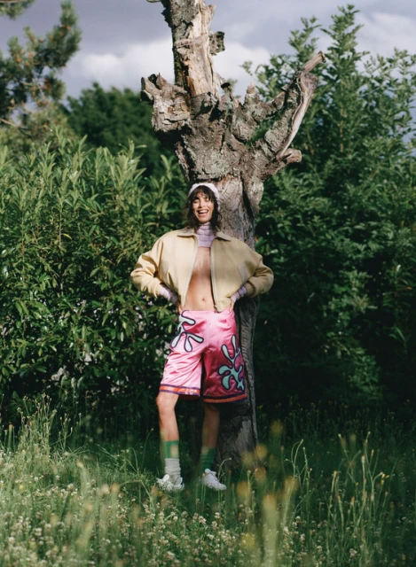 Adult posing in pink patterned shorts and tan jacket next to a tree in a grassy field with green trees and cloudy skies.