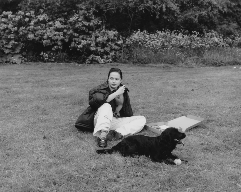 Young woman sits in a grassy field with her black dog, sharing pizza on a monochromatic day, with leafy bushes in the background.