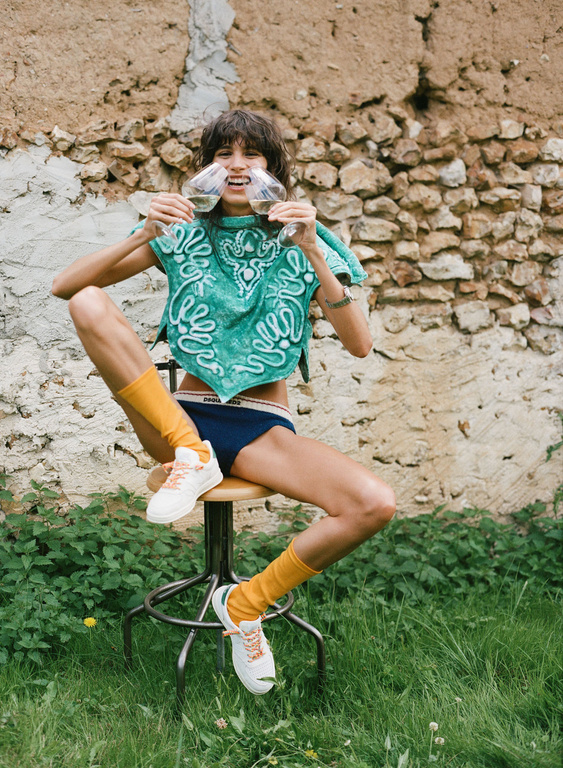 Woman in teal top and underwear sits on stool outside holding wine glasses, wearing yellow socks and white sneakers near stone wall.