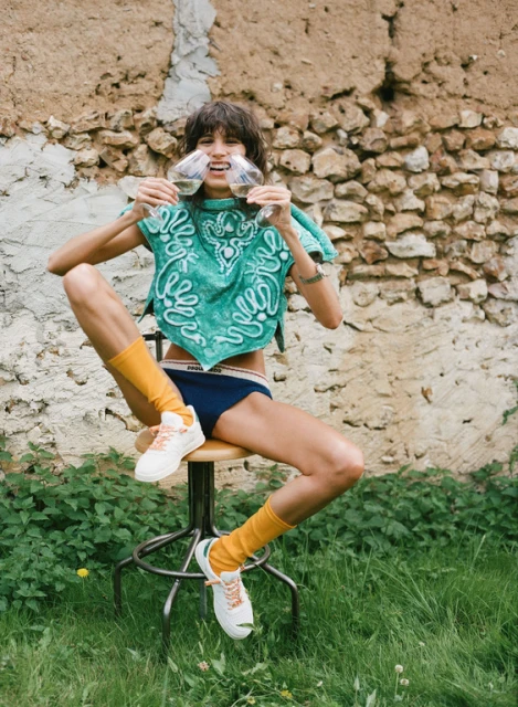 Woman in teal top and underwear sits on stool outside holding wine glasses, wearing yellow socks and white sneakers near stone wall.