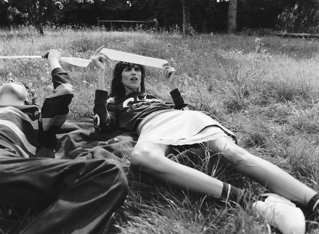 Two people lying on grass outdoors, holding up a light-colored ribbon in a park setting.