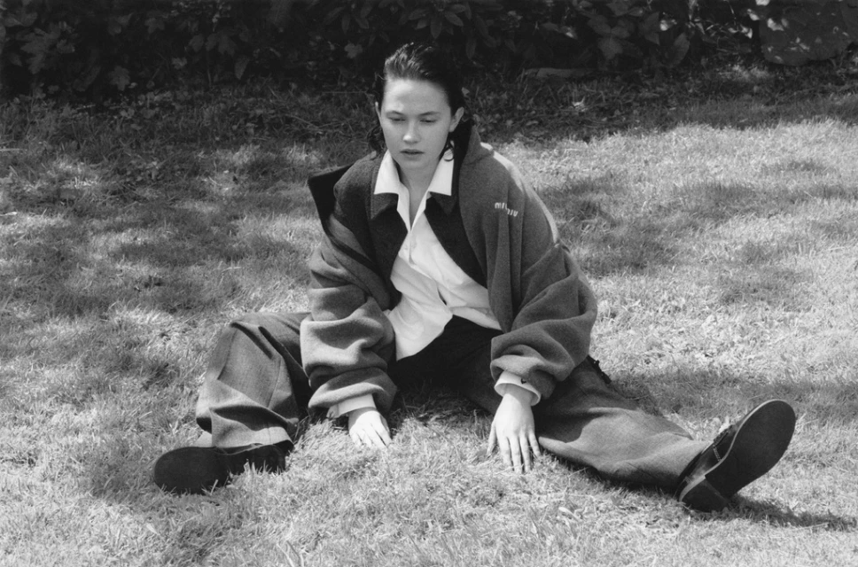 Woman sits in grass wearing oversized jacket and pants in a black and white outdoor portrait, casual fashion aesthetic.