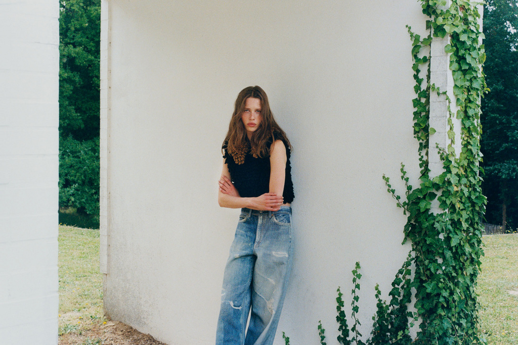 A young woman with long brown hair posing in front of a white wall, wearing a dark top and light blue jeans in an outdoor setting.