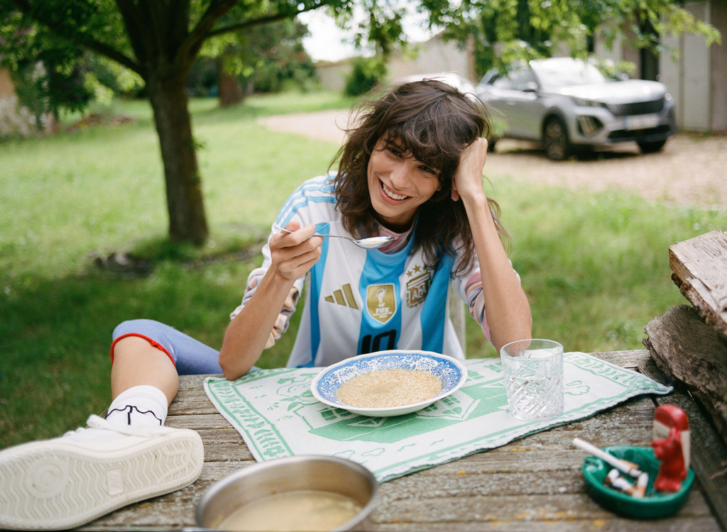 Woman in Argentina soccer jersey eating outdoors at a wooden table with food, a glass of water, and a car in the background.