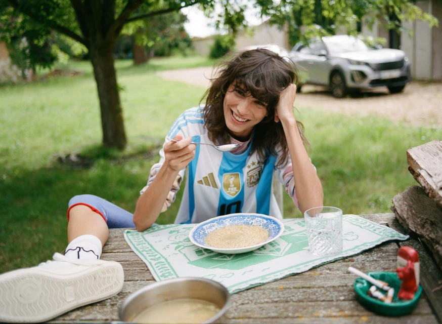 Woman in Argentina soccer jersey eating outdoors at a wooden table with food, a glass of water, and a car in the background.