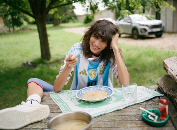 Woman in Argentina soccer jersey eating outdoors at a wooden table with food, a glass of water, and a car in the background.