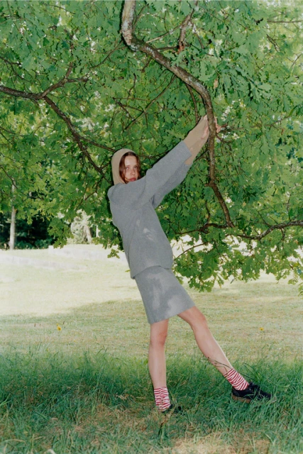 A person poses in a gray suit and striped socks, leaning against a tree in a grassy field under natural light, creating a whimsical outdoor portrait.