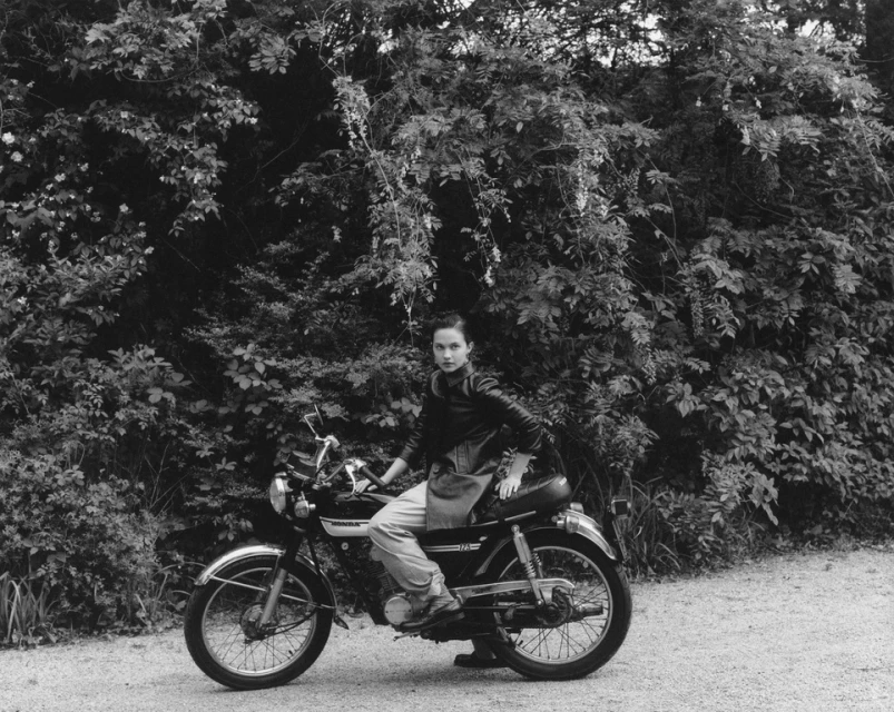 Woman sits on a vintage motorcycle on a dirt road in a natural setting, surrounded by foliage. Black and white photo.