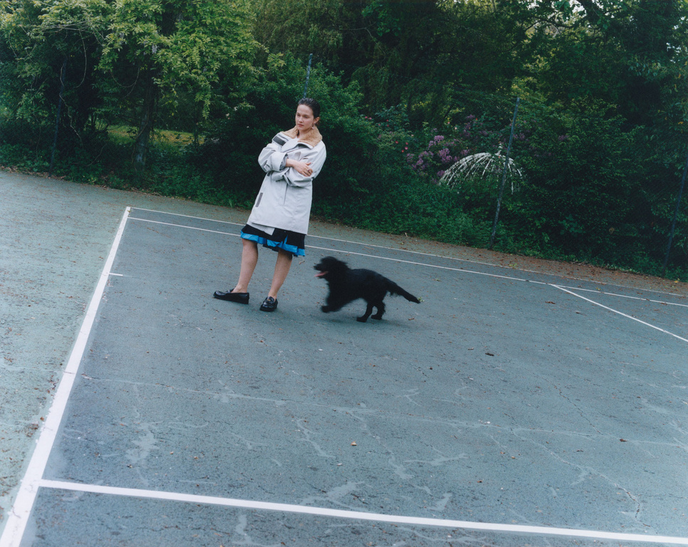 A woman stands on a tennis court with a black dog running nearby, surrounded by lush greenery, wearing a beige coat and dark skirt.