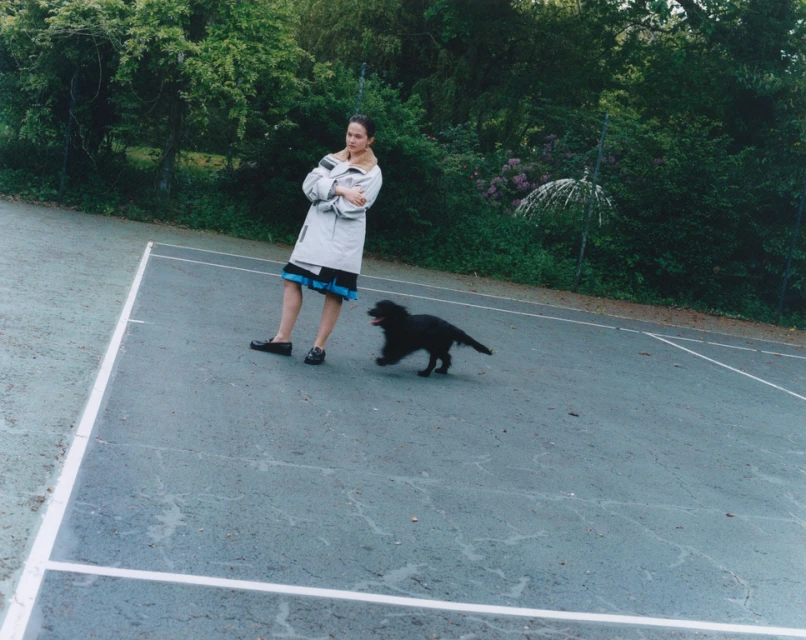A woman stands on a tennis court with a black dog running nearby, surrounded by lush greenery, wearing a beige coat and dark skirt.