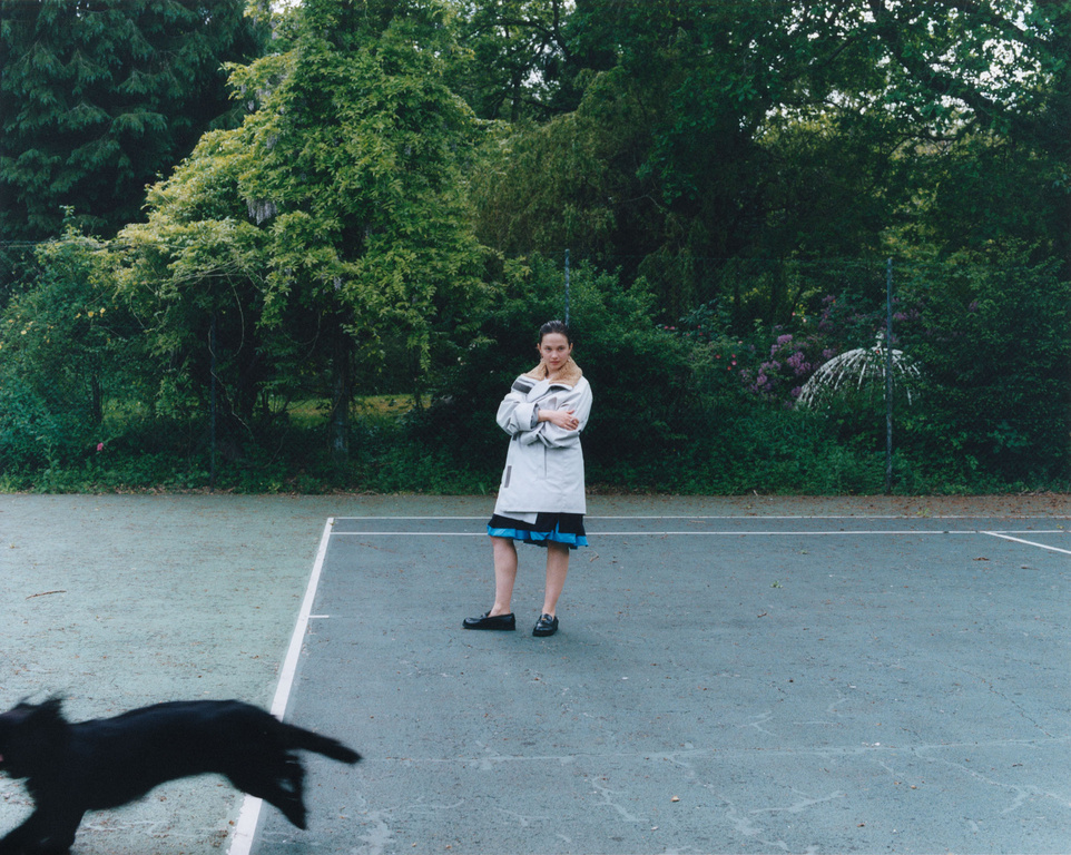 A woman stands on a tennis court wearing a coat and skirt as a black dog runs past, with trees and foliage in the background on an overcast day.