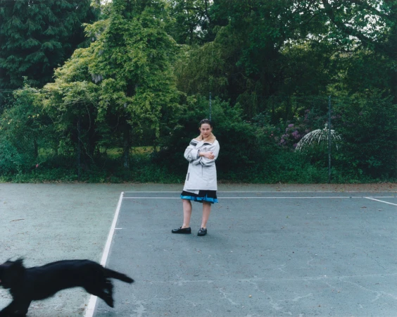 A woman stands on a tennis court wearing a coat and skirt as a black dog runs past, with trees and foliage in the background on an overcast day.
