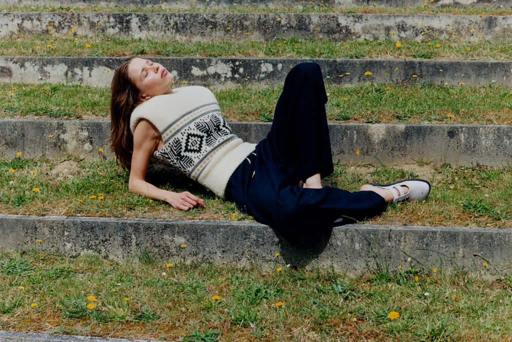 A young woman with long brown hair lies on concrete steps in a sleeveless sweater and dark pants enjoying the sunlight.