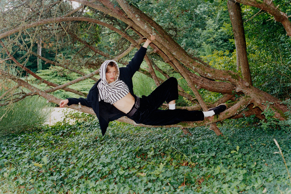 Young adult woman in casual fashion perched in a tree with green foliage background, wearing a scarf, crop top, and black jacket.