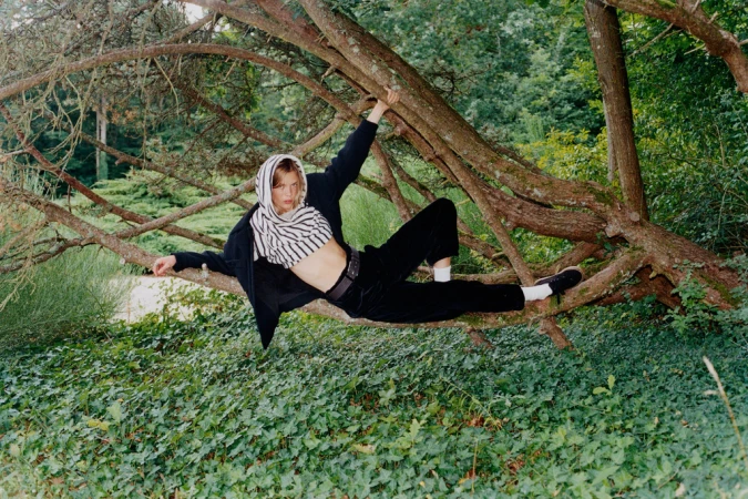 Model in striped hooded top and dark trousers climbs amidst thick tree branches over a lush green woodland floor.