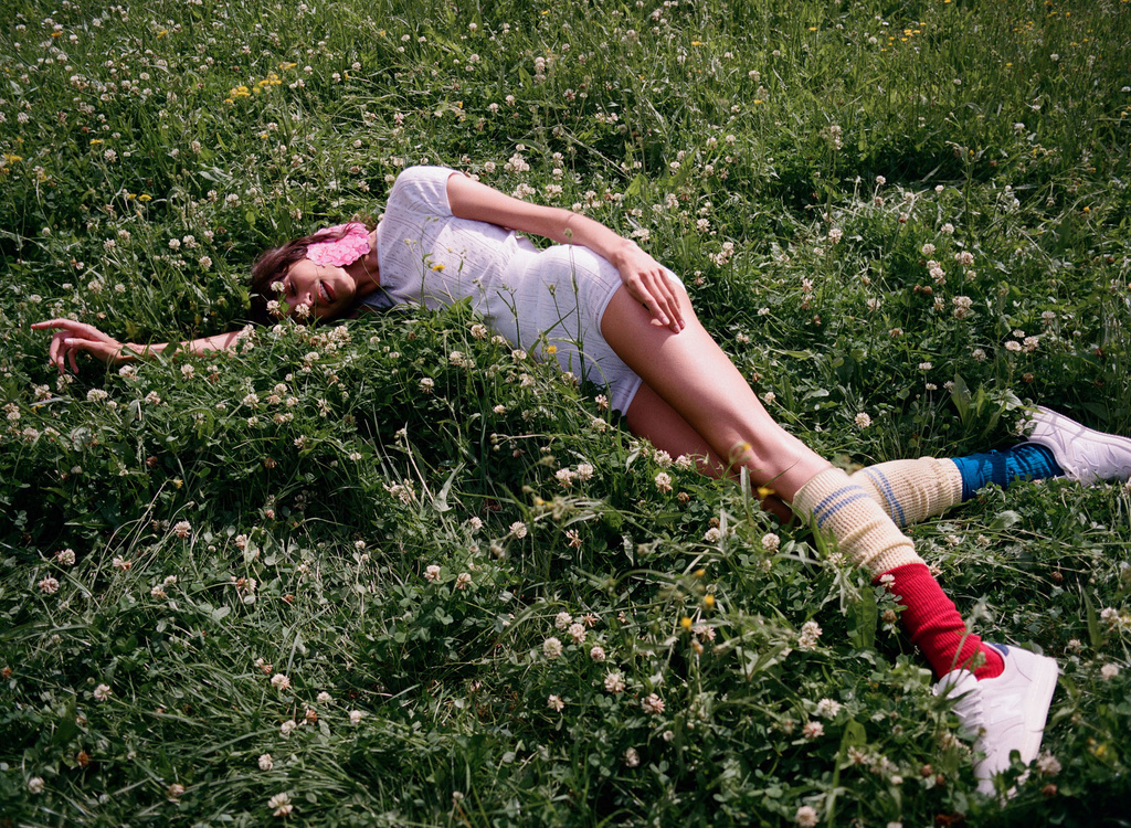 Woman in a white romper lies in a field of clovers, wearing colorful socks and white sneakers in an outdoor setting.