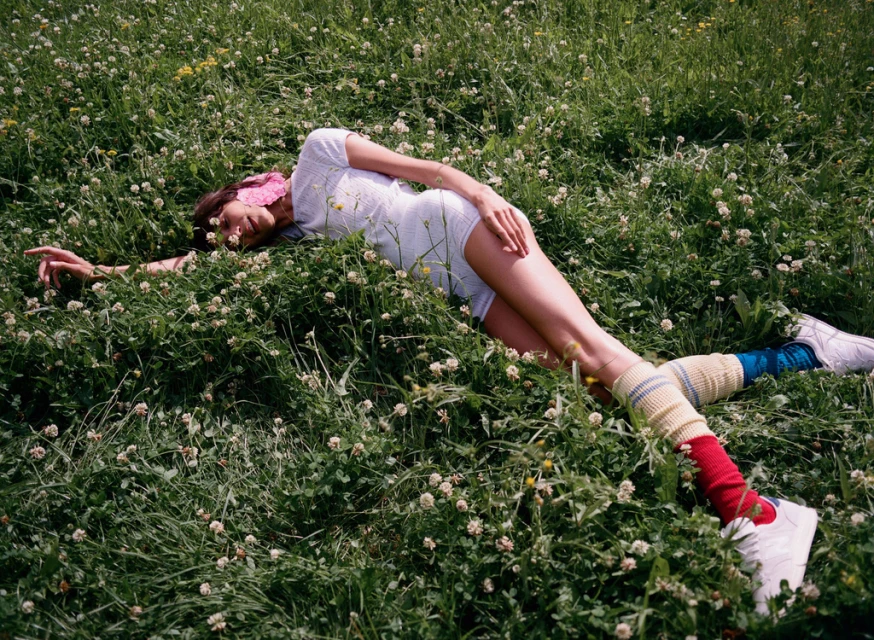 Woman in a white romper lies in a field of clovers, wearing colorful socks and white sneakers in an outdoor setting.