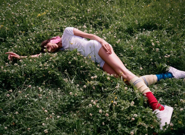 Woman in a white romper lies in a field of clovers, wearing colorful socks and white sneakers in an outdoor setting.