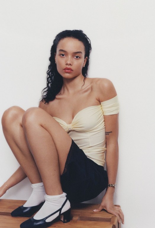 Young adult woman poses seated indoors wearing a pale yellow off-the-shoulder top, dark mini skirt, white socks, and black heeled shoes.