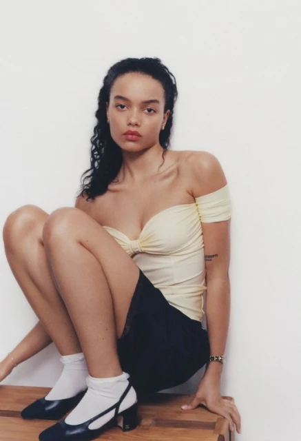 Young adult woman poses seated indoors wearing a pale yellow off-the-shoulder top, dark mini skirt, white socks, and black heeled shoes.