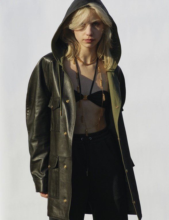 A young blonde woman poses in an olive-green hooded jacket and black bikini top against a bright white background in a fashion style portrait.