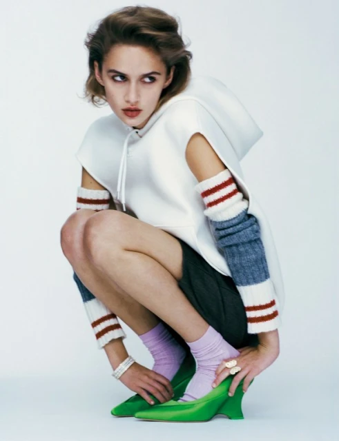 Fashion shot of a fair-skinned woman crouching in green heels, lilac socks, a white hoodie, and gray shorts against a plain background.