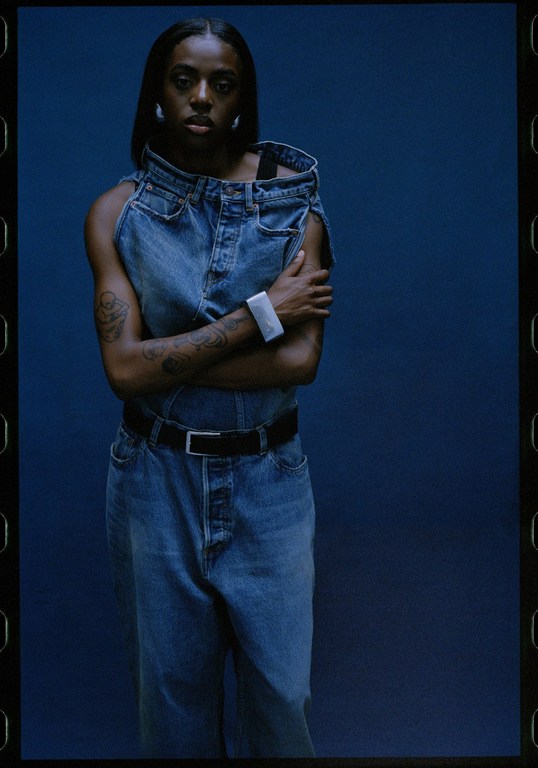 A young Black woman stands in front of a blue backdrop, modeling a denim outfit and accessorized with hoop earrings and a smart watch.