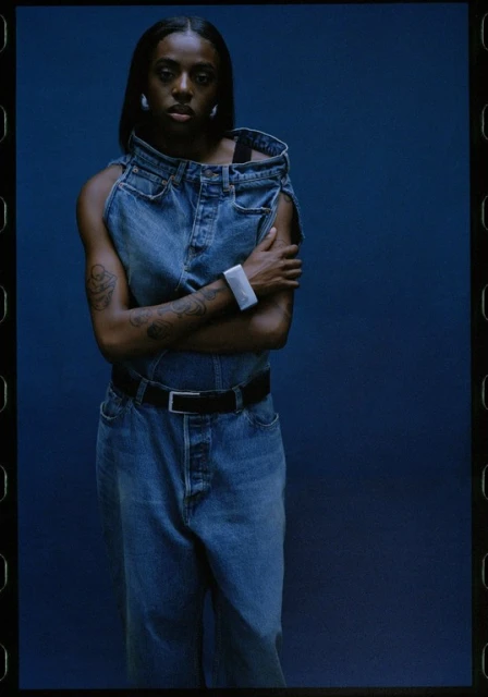 A young Black woman stands in front of a blue backdrop, modeling a denim outfit and accessorized with hoop earrings and a smart watch.
